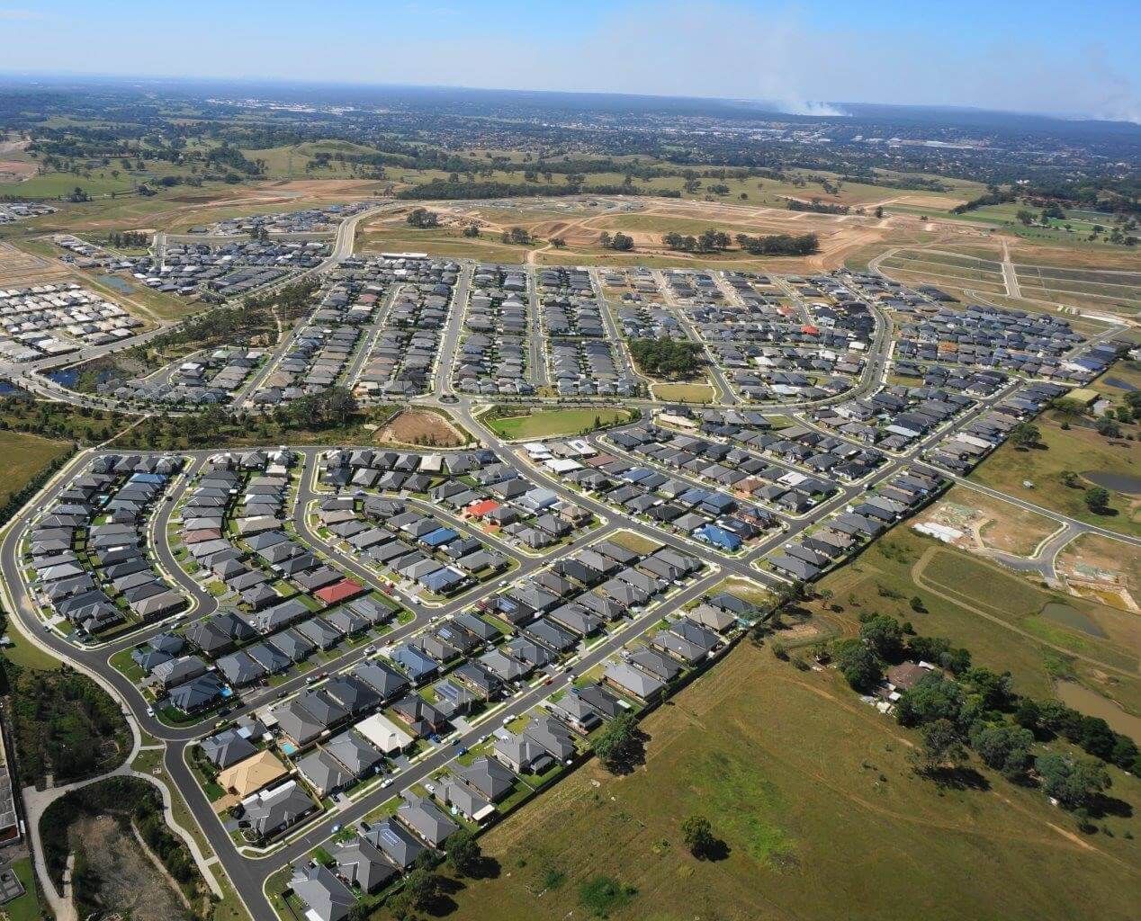 Aerial view of a suburban residential area with rows of houses, roads, and patches of green space under a clear blue sky.