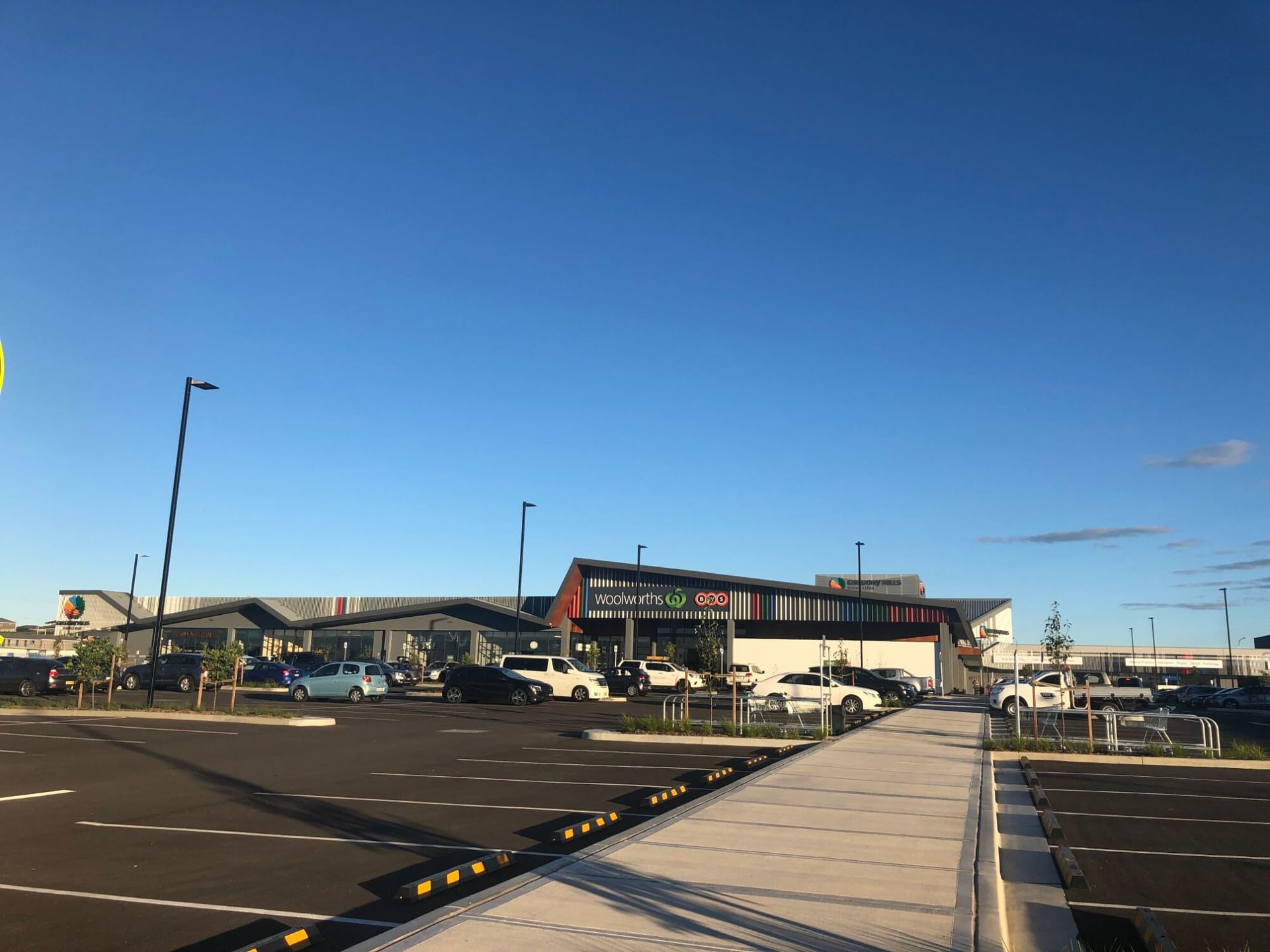 Empty parking lot with a shopping center in the background under a clear blue sky. Cars are parked around the lot.