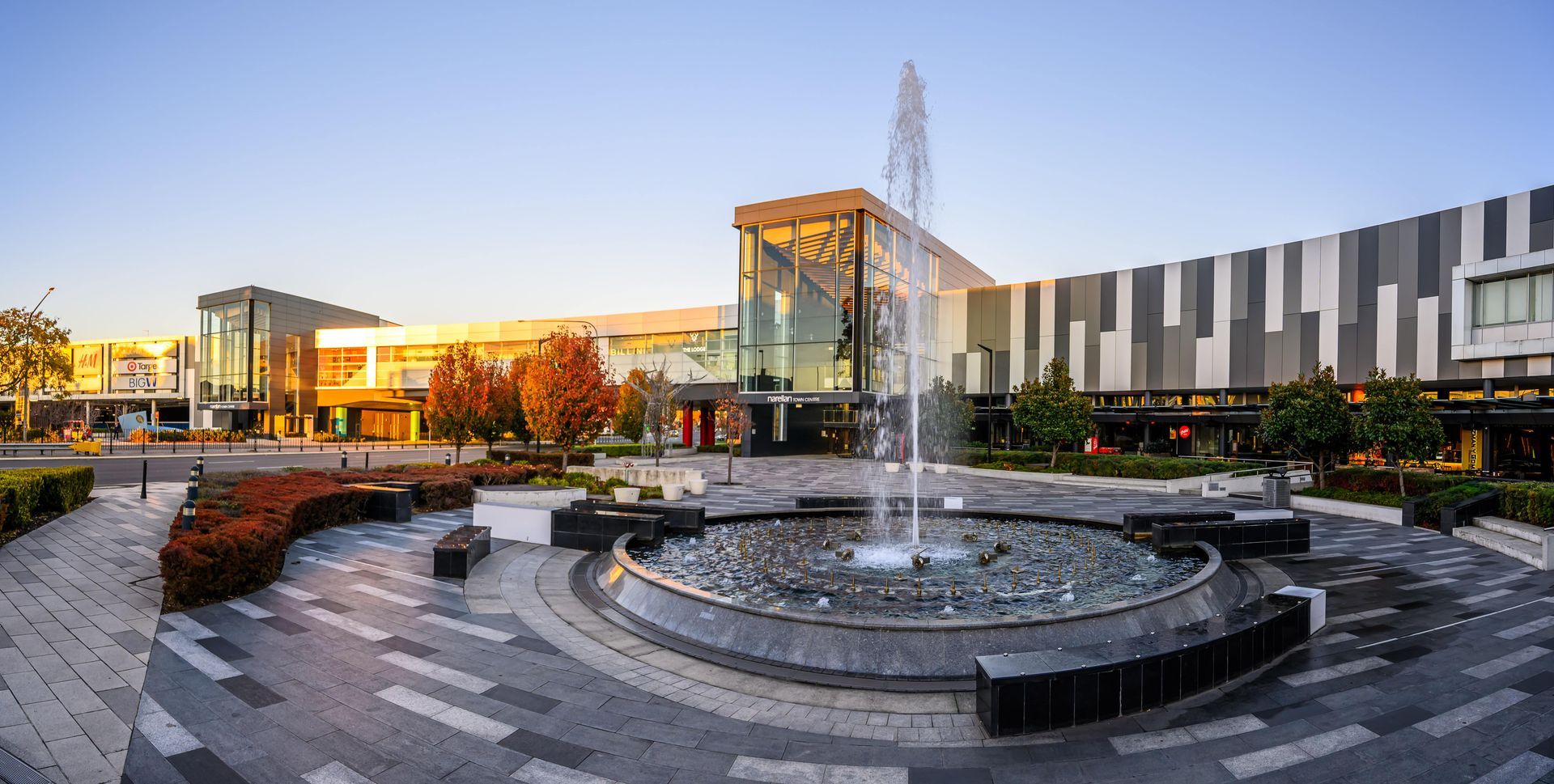 Exterior view of a modern shopping mall at dusk, featuring a fountain in the foreground and contemporary architecture.