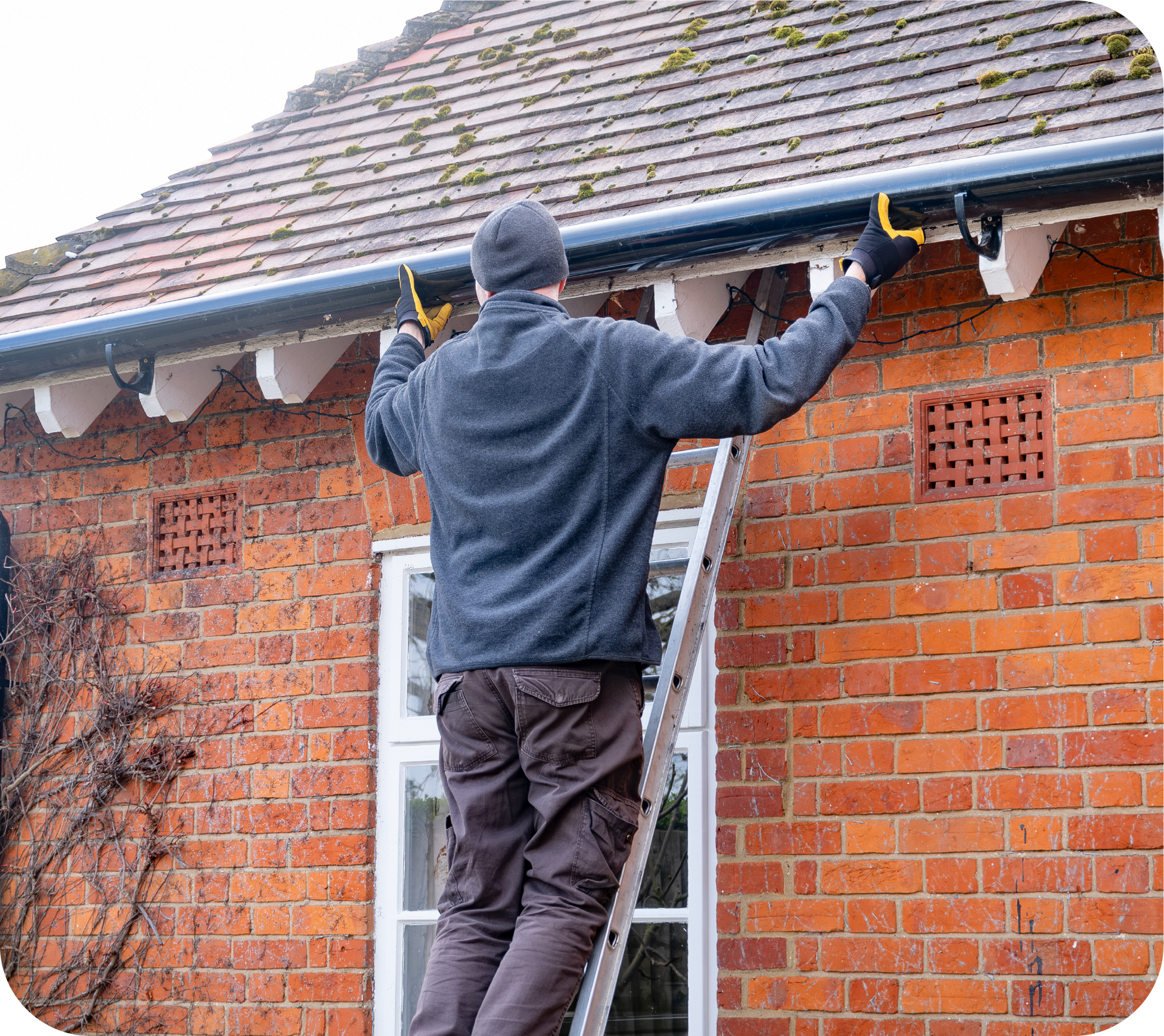A man is standing on a ladder fixing a gutter on a brick building.
