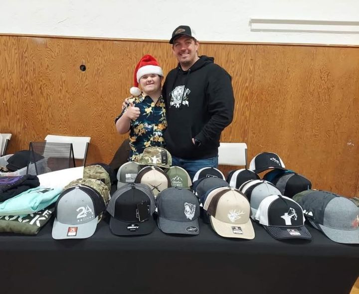 Two people behind a table of baseball caps, posing at an indoor vendor booth