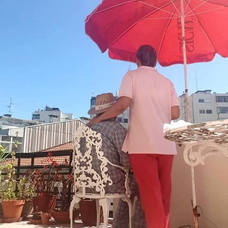 Uma mulher de camisa rosa está ao lado de um homem em uma cadeira sob um guarda-chuva vermelho