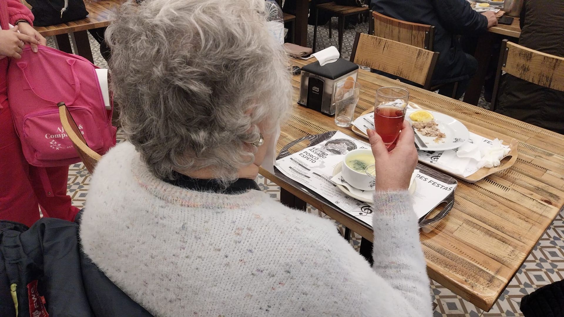 Uma senhora idosa está sentada à mesa de um restaurante segurando uma bebida.