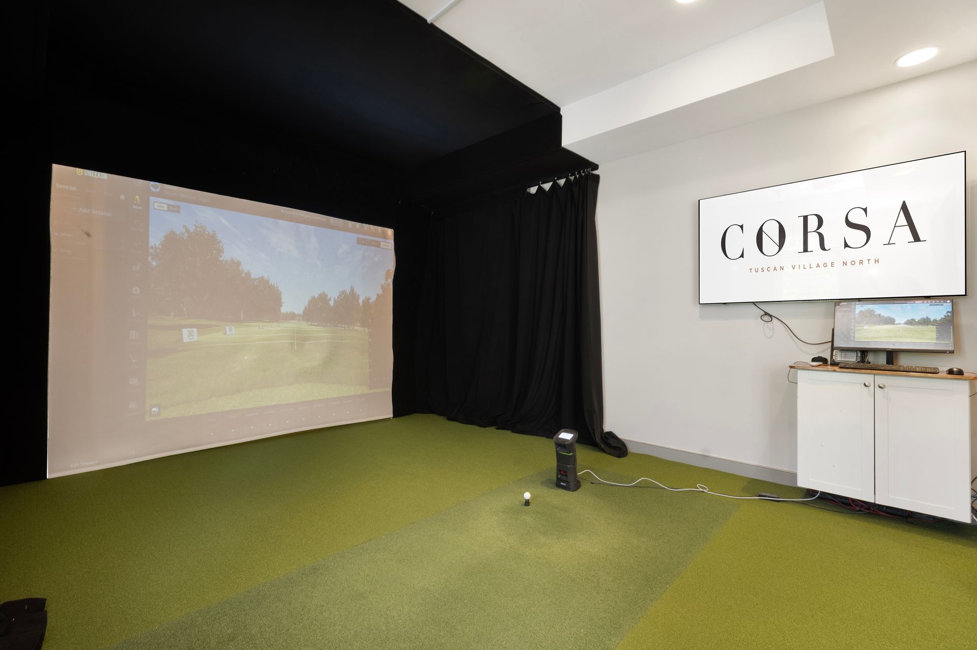Indoor golf simulator room with projector screen, green turf, and CORSA sign on the wall at Corsa Tuscan Village in Salem, NH.
