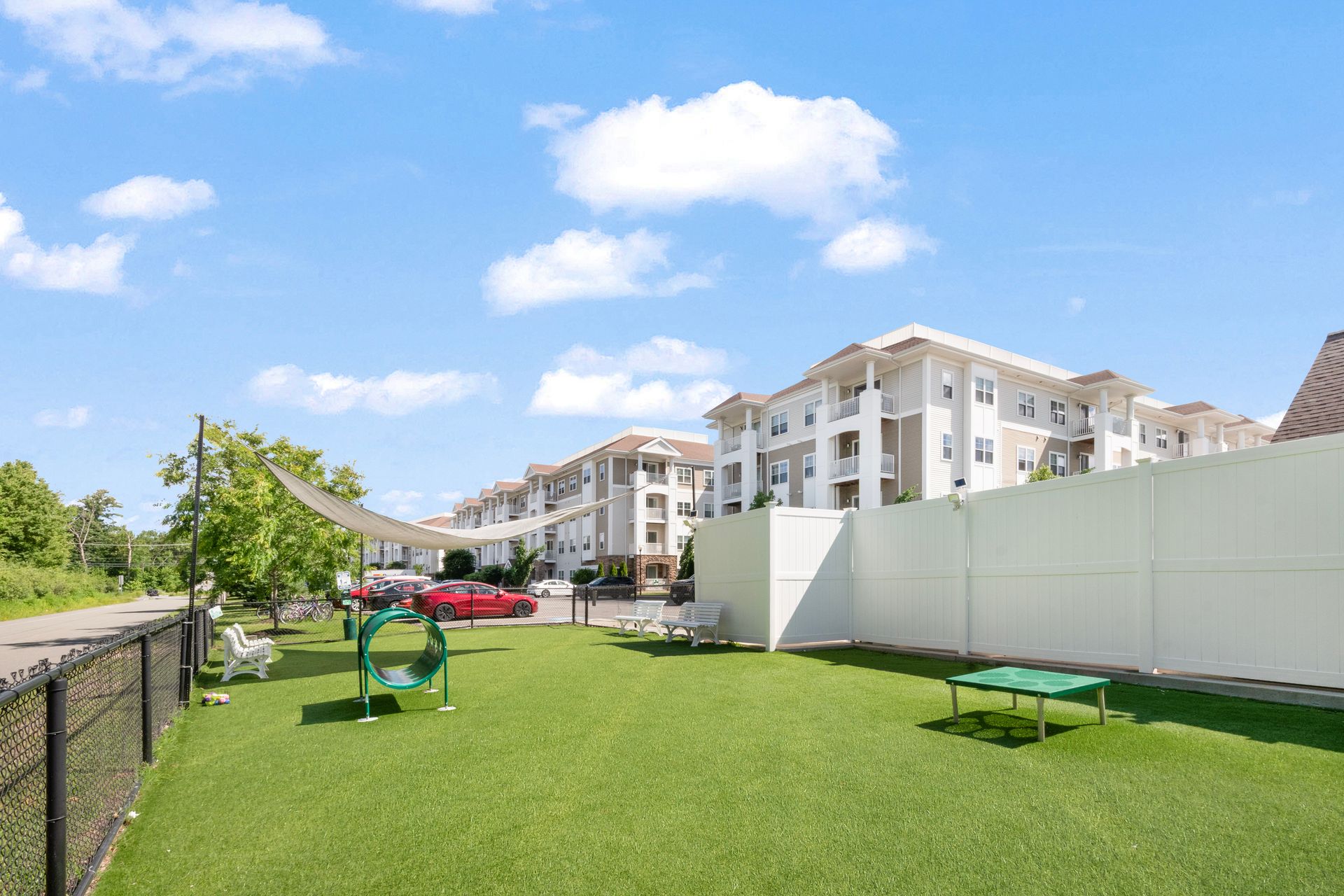 Sunny apartment courtyard with green lawn, benches, playground equipment, and white residential buildings at Corsa Tuscan Village in Salem, NH.