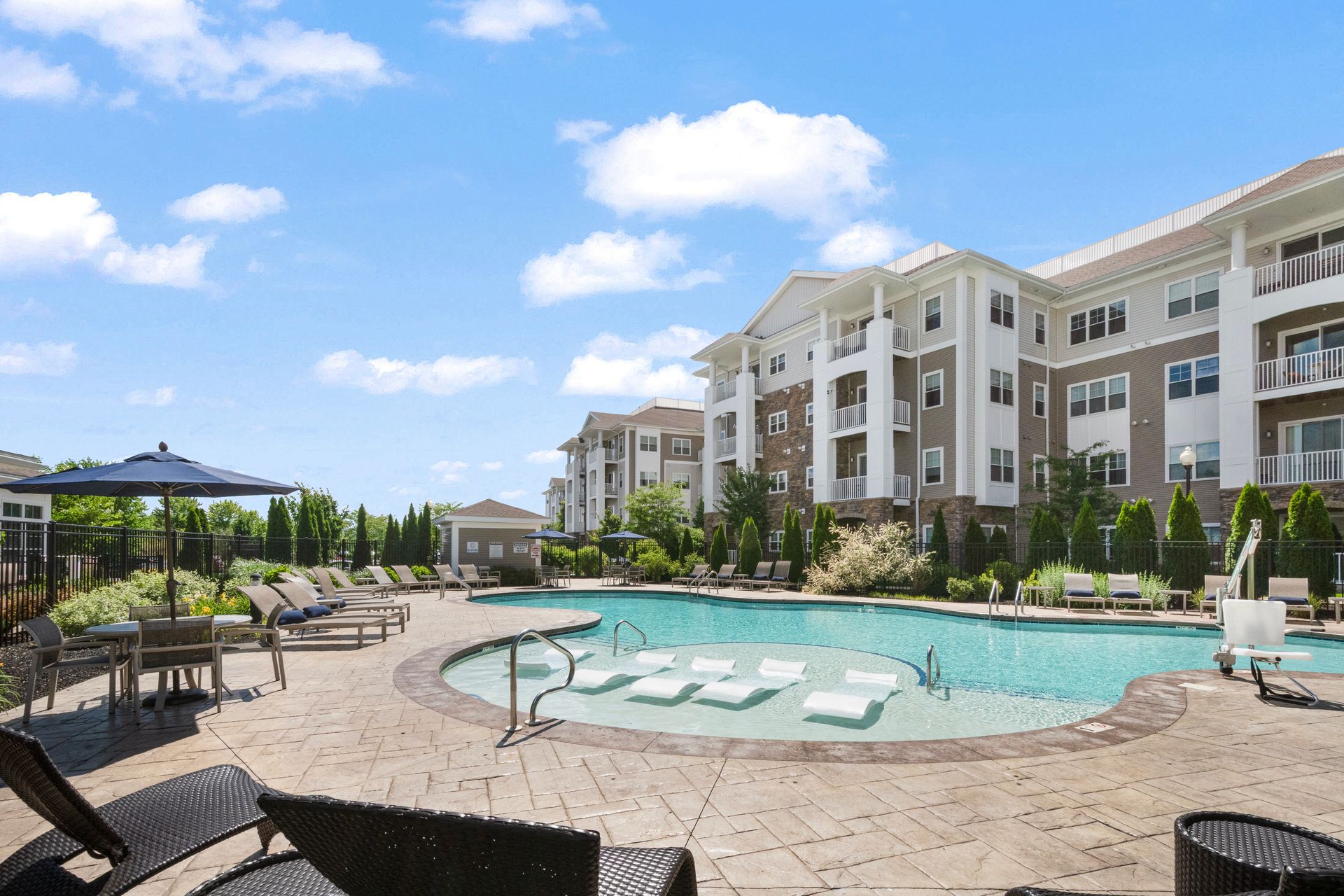 Resort pool with lounge chairs and multi-story buildings under a bright blue sky at Corsa Tuscan Village in Salem, NH.
