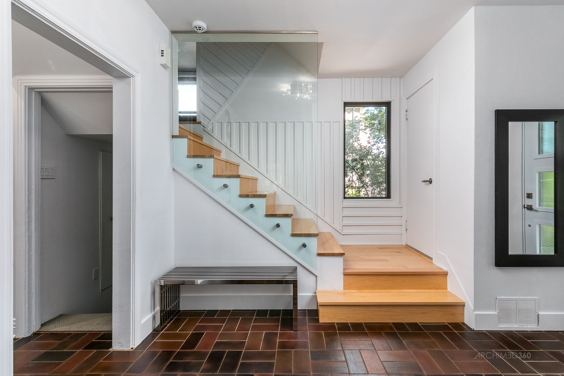 Un couloir avec des escaliers, un banc, un miroir et une fenêtre.