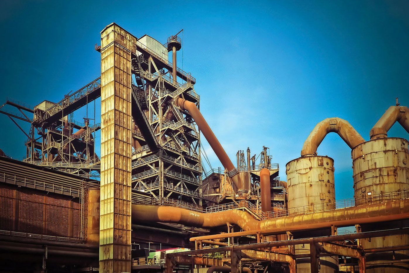 Rusty industrial steel mill against a blue sky, with pipes and structures.