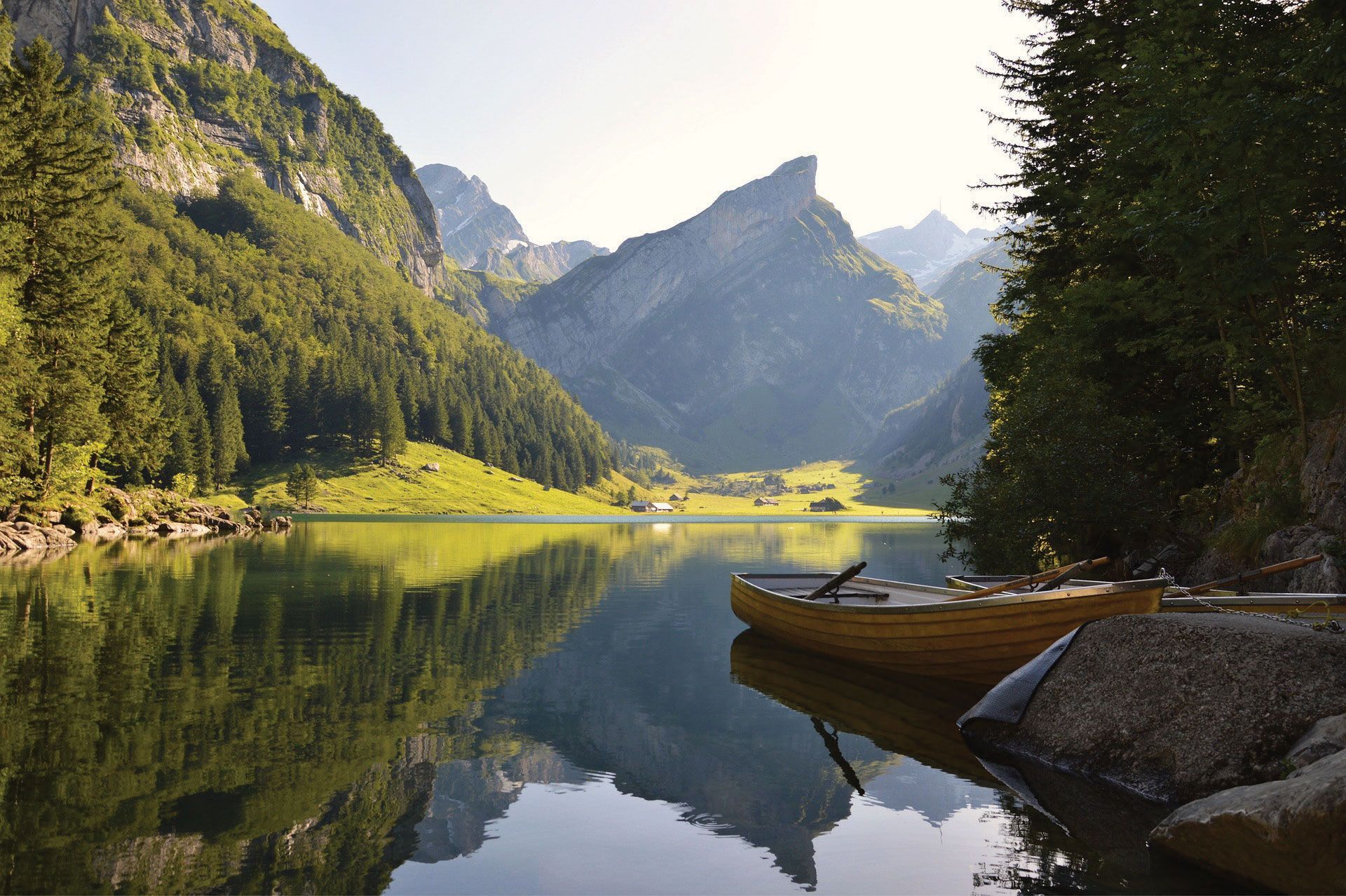 Wooden rowboat on calm lake, mountains reflected in water; lush green forest and meadow.