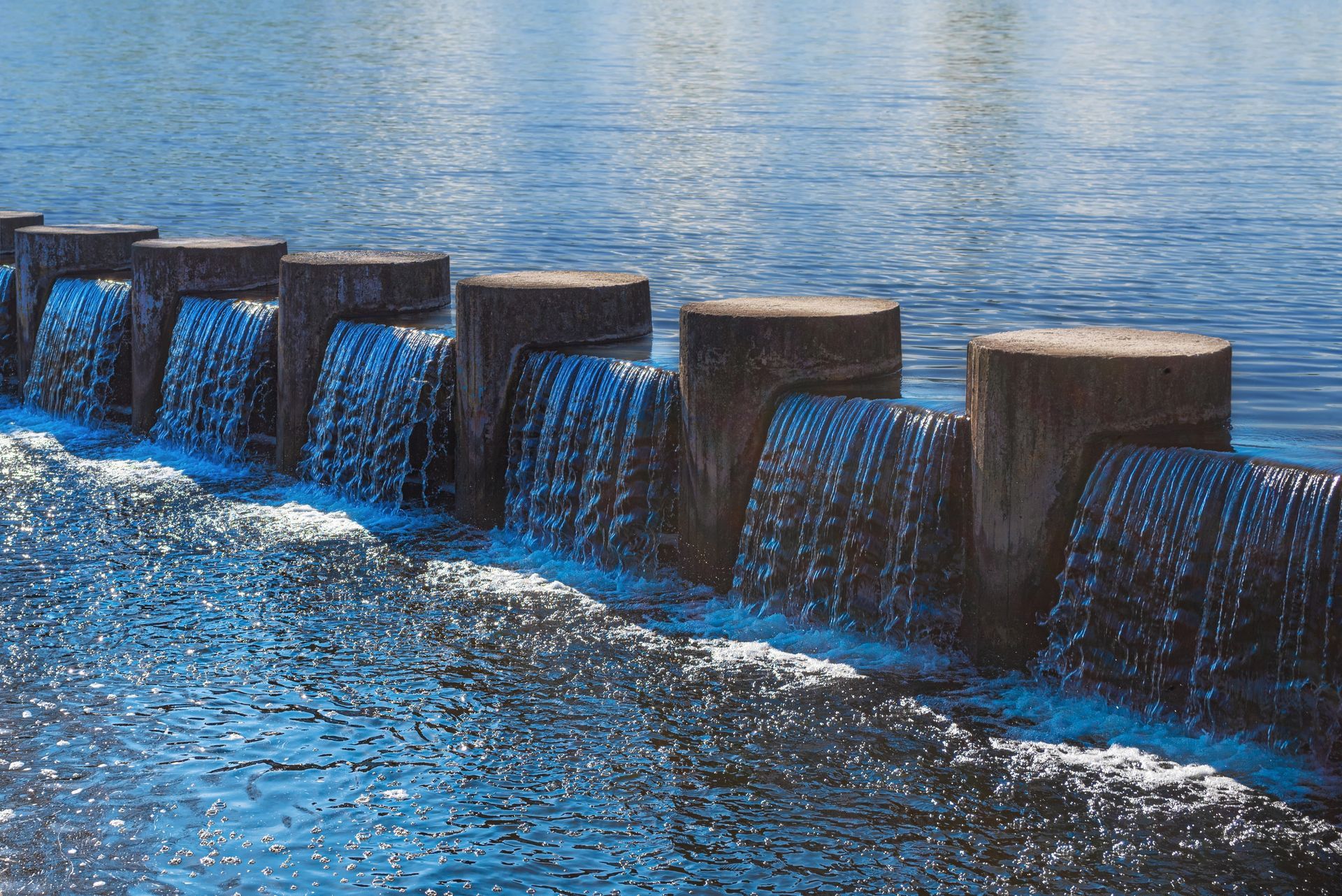A concrete dam with water cascading over its top into a blue river