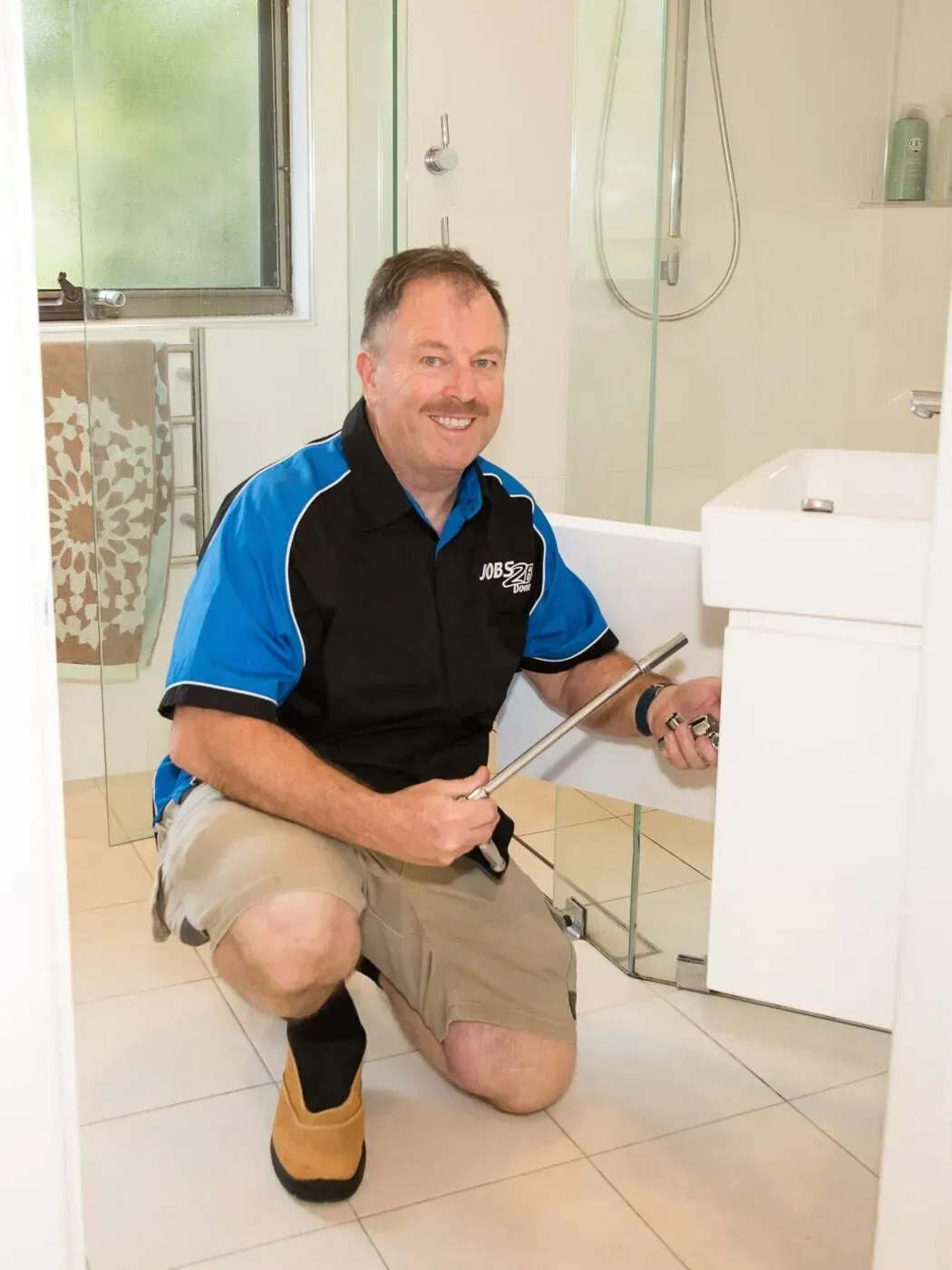 A man is kneeling down in a bathroom next to a sink. — Jobs2BDone in Woy Woy, NSW