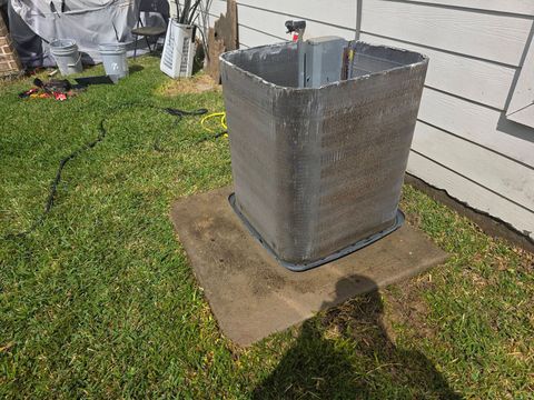 Square metal outdoor unit on a concrete pad beside a house, with grass and a hose nearby.