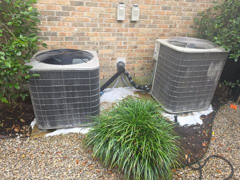 Two outdoor HVAC units by a brick wall, with snow on top and gravel and plants around them.