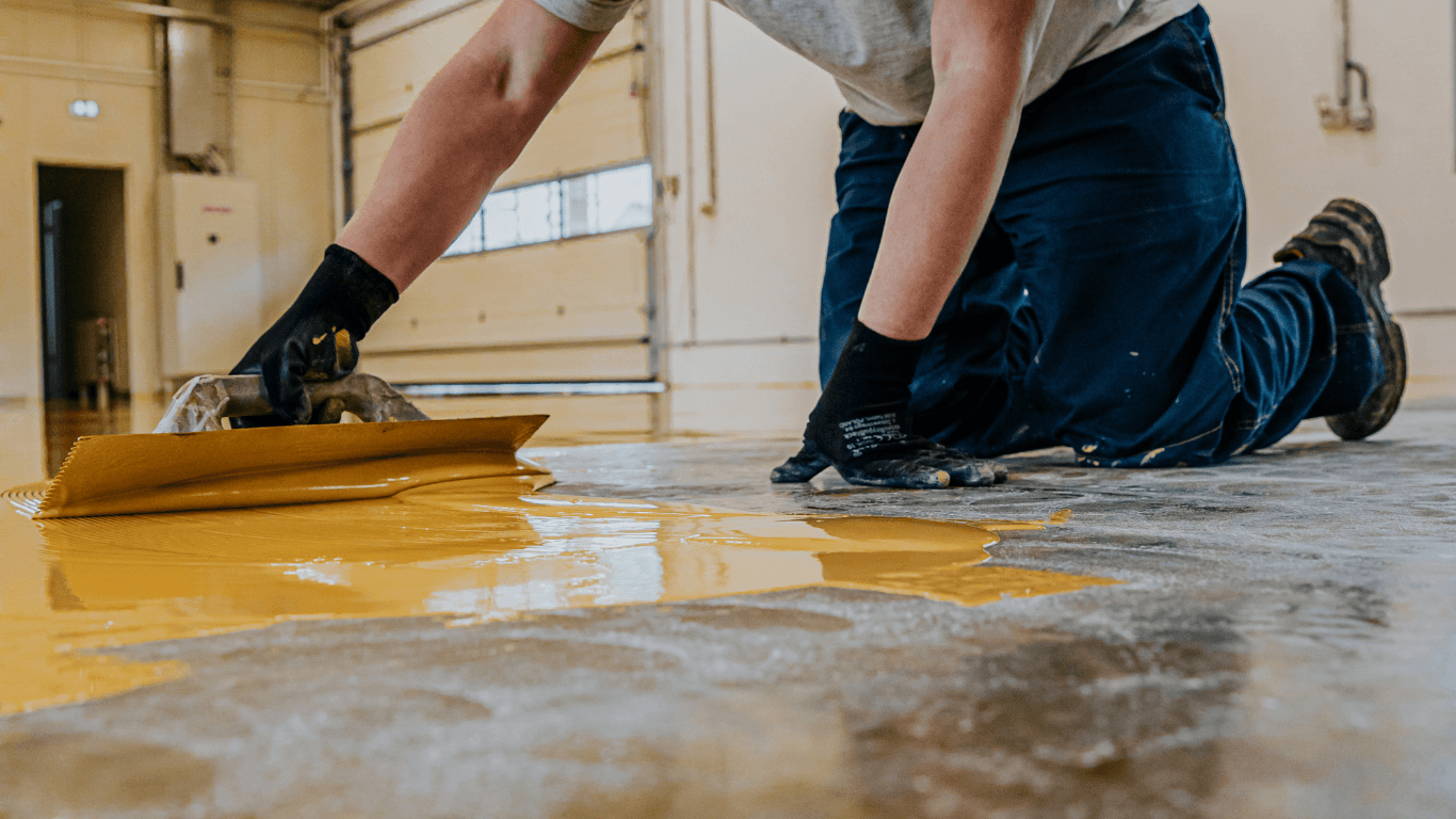 Worker kneeling and spreading epoxy coating across a floor with a tool to ensure even coverage during an epoxy flooring service in Glendale, AZ