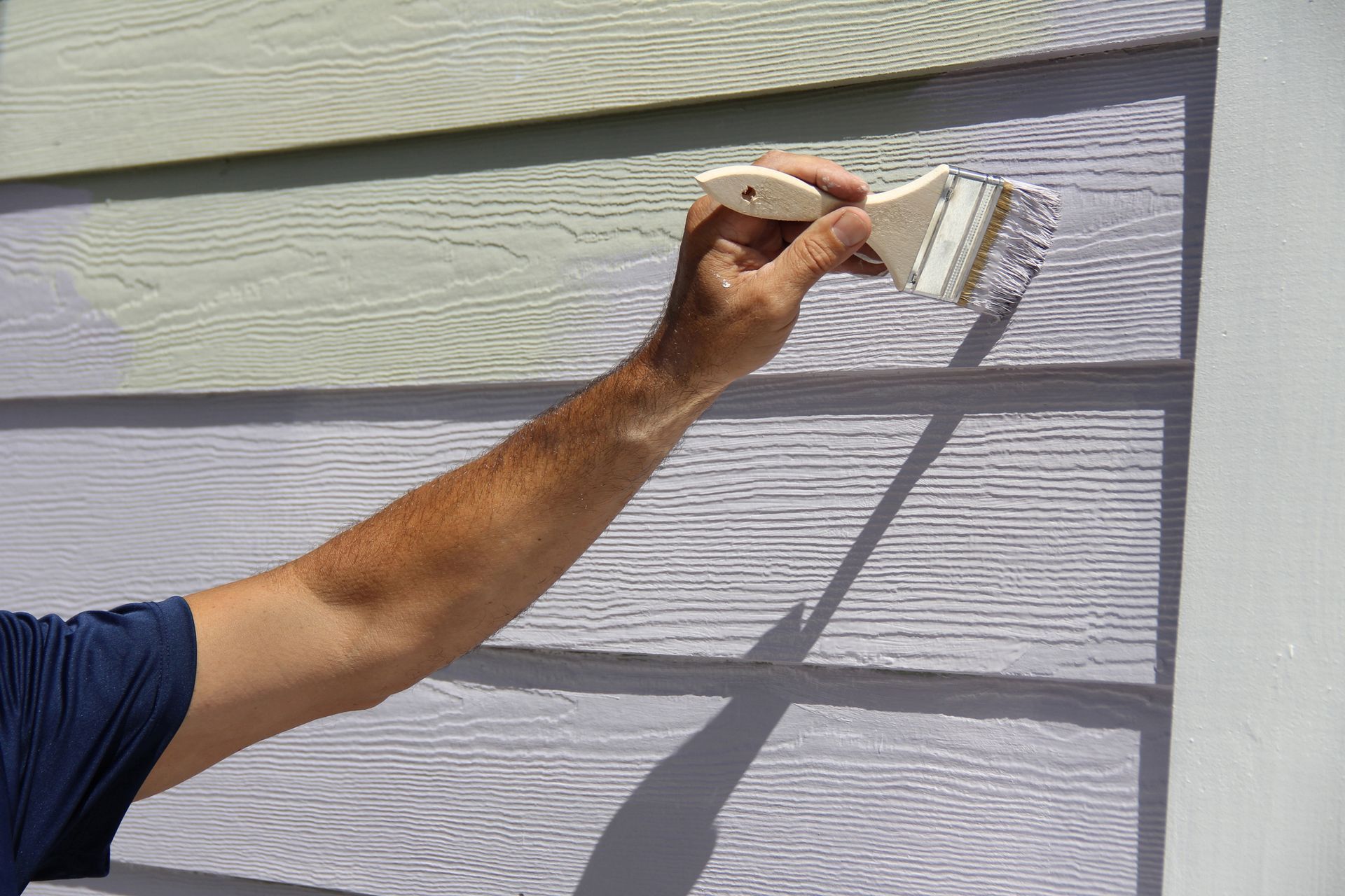 Person painting light-colored siding with a paintbrush, outdoors.