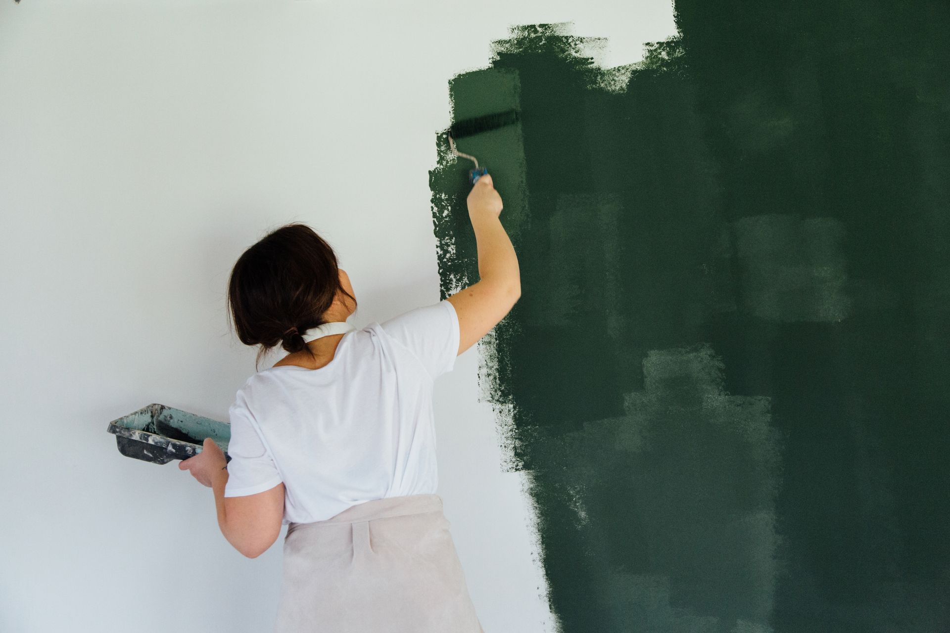Person painting a wall dark green with a roller. White shirt and apron.