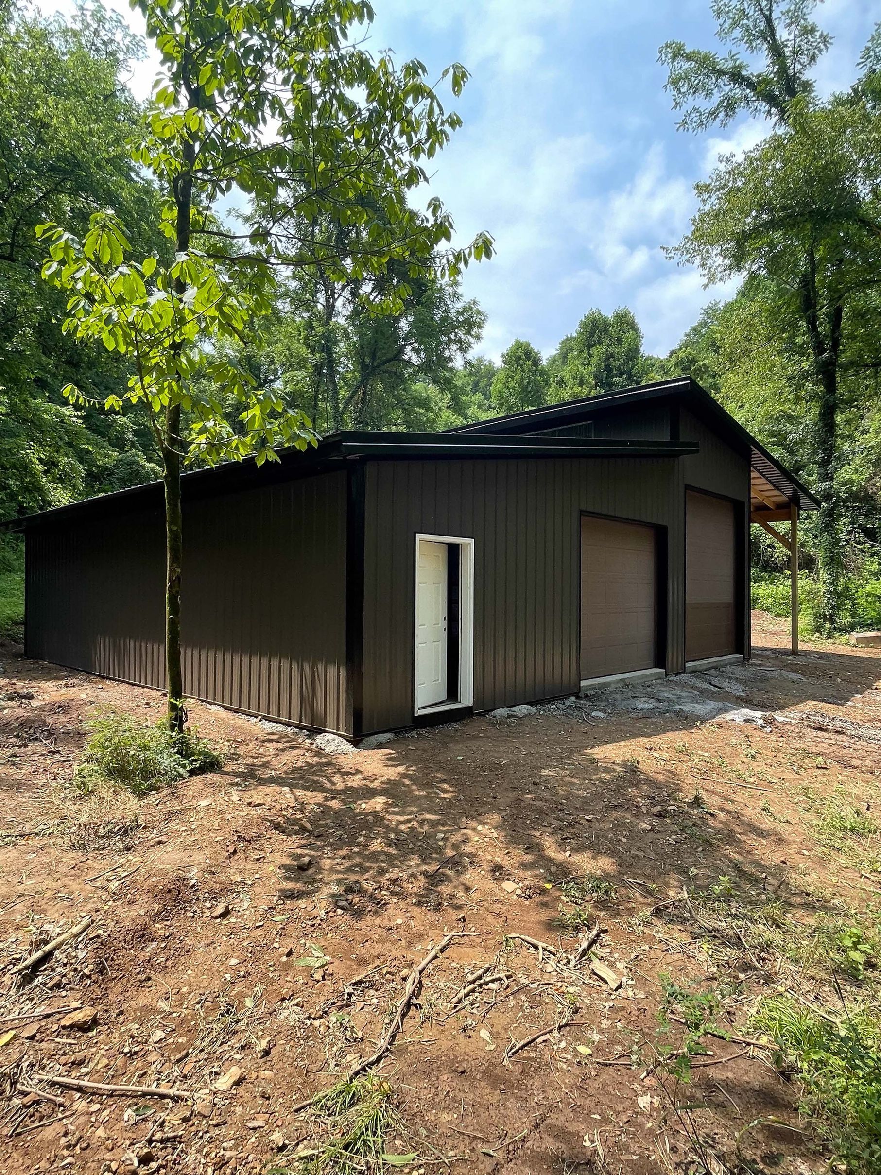 A large black garage is sitting in the middle of a dirt field surrounded by trees.