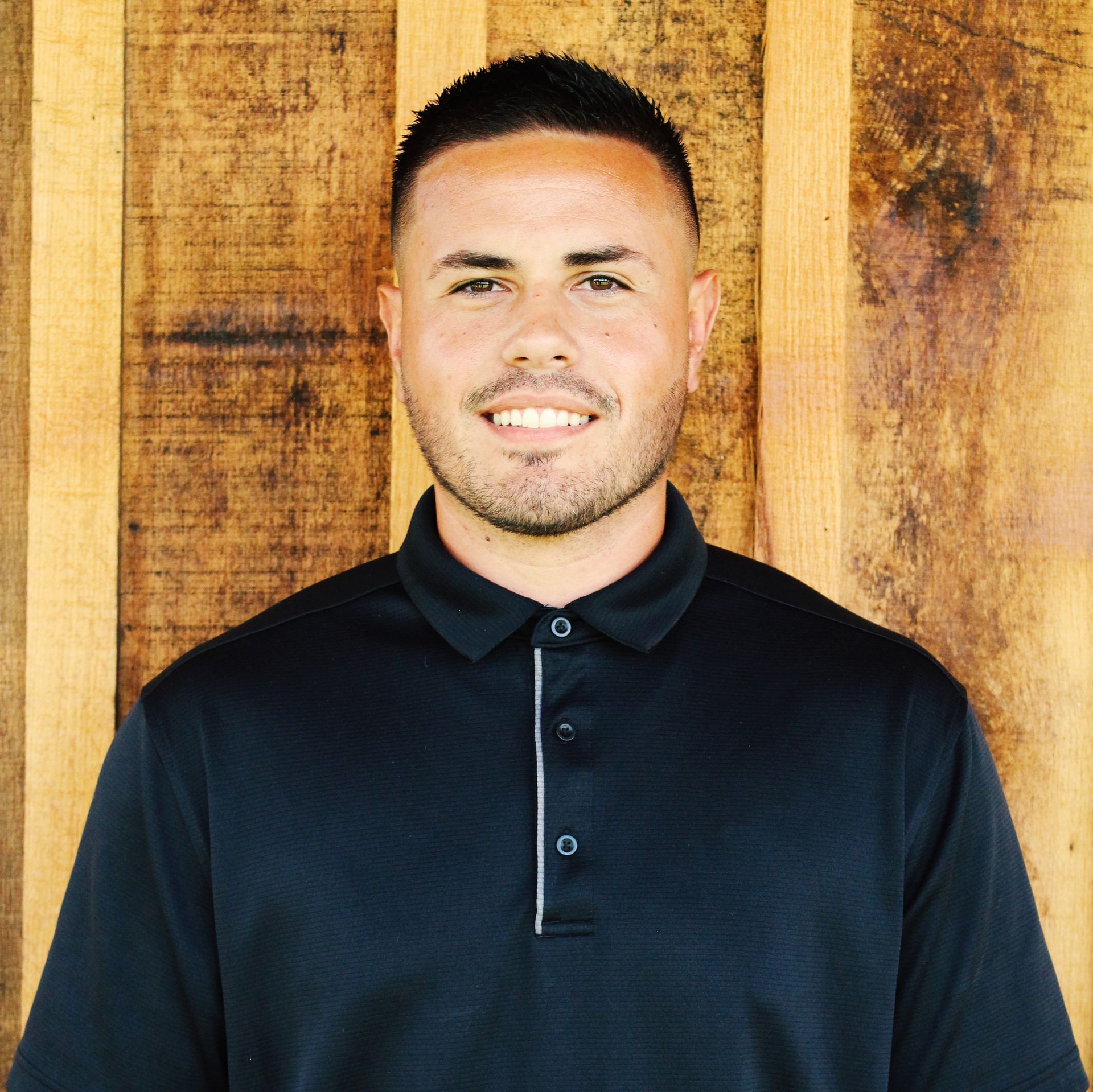 A man in a black polo shirt is smiling in front of a wooden wall