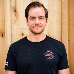 A man wearing a black t-shirt with a logo on it is standing in front of a wooden wall.