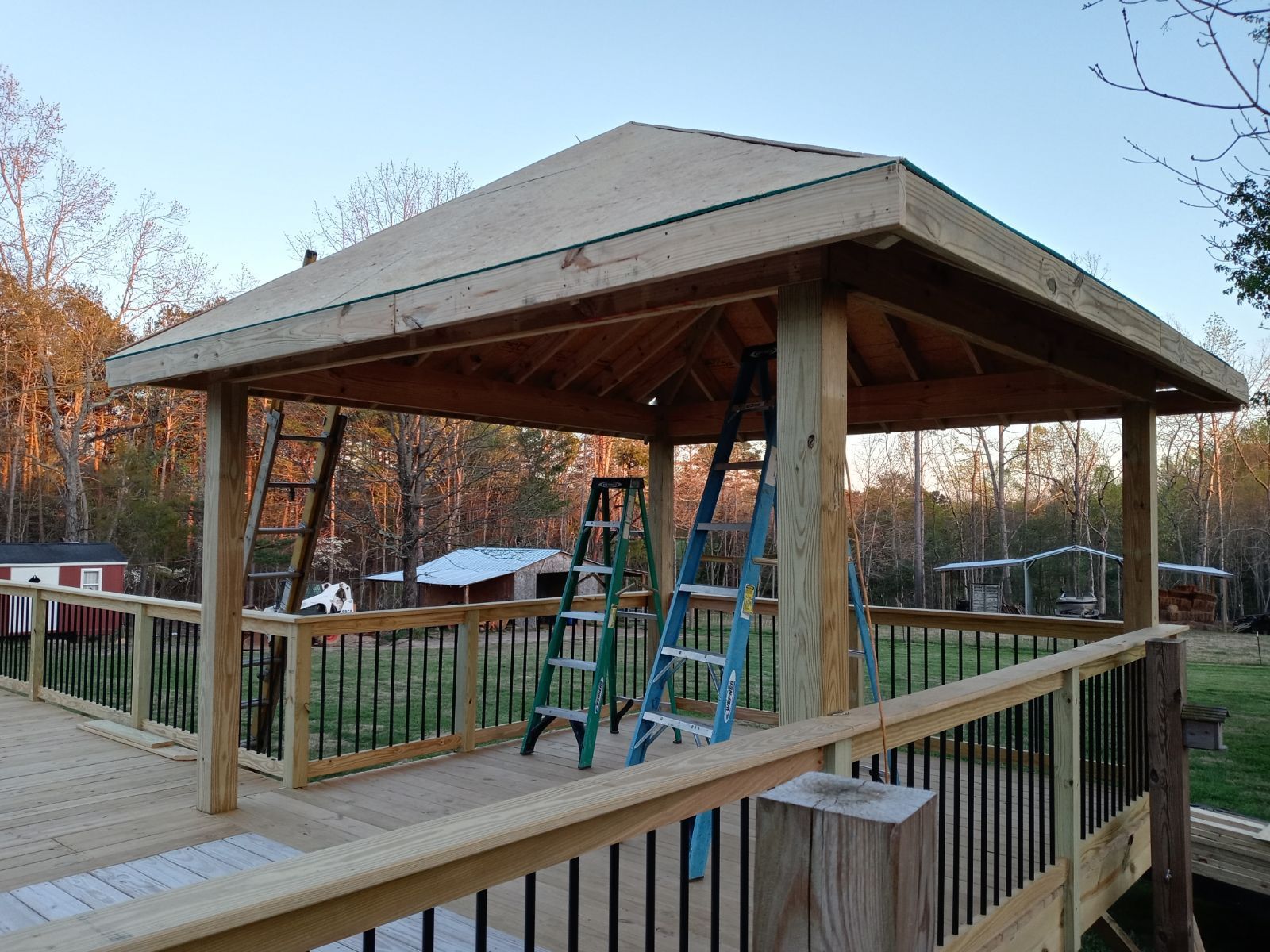 Wooden gazebo under construction on a deck, with ladders inside and a backyard setting.