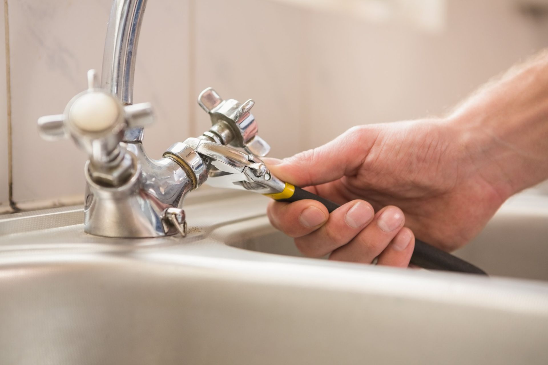A hand using a wrench on a chrome faucet in a kitchen sink.