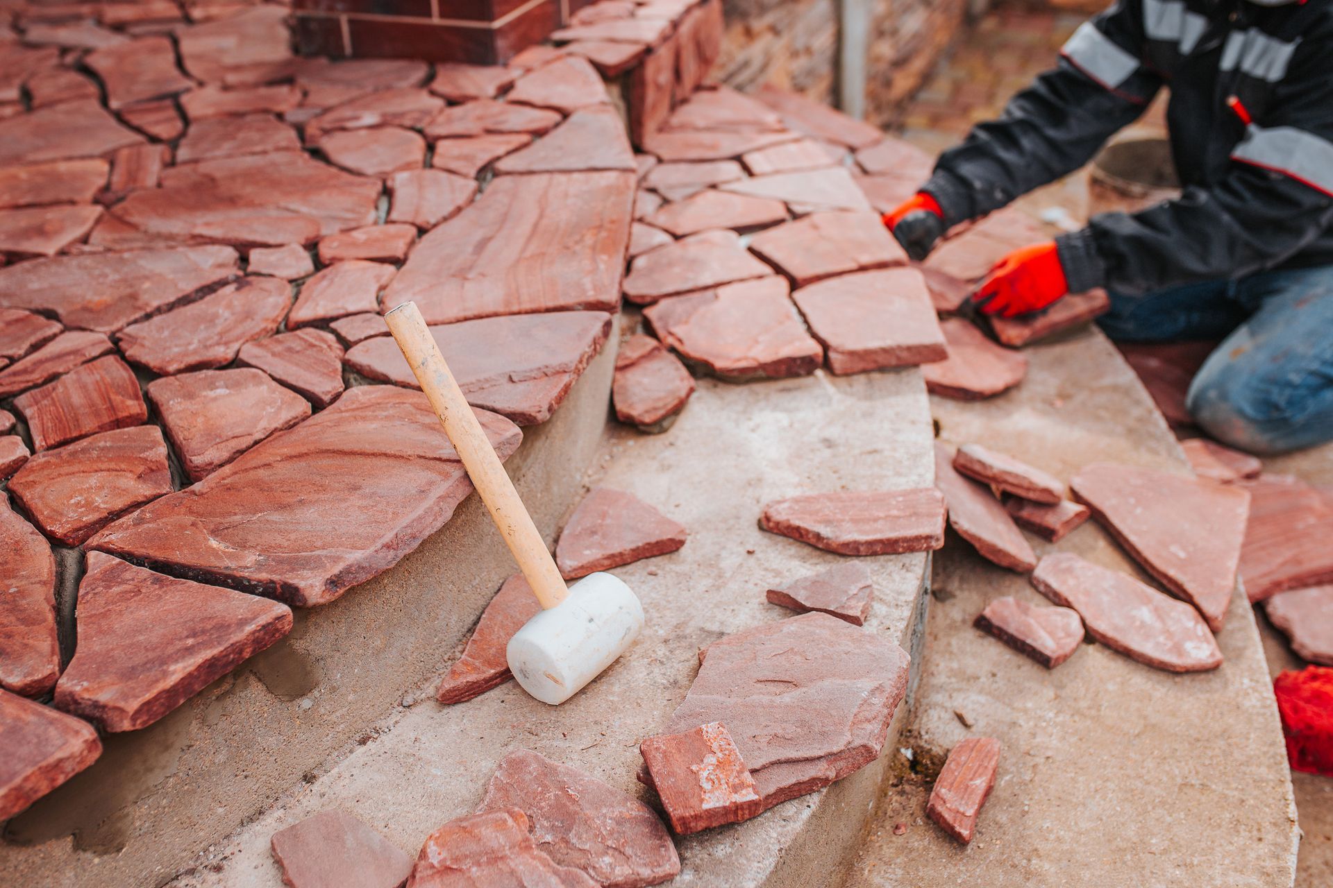 Man laying reddish-brown stone tiles on steps, using a mallet, outdoors.