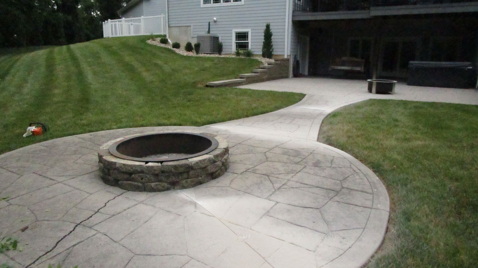 Concrete patio with fire pit in a backyard, leading to a house. Green lawn and trees.