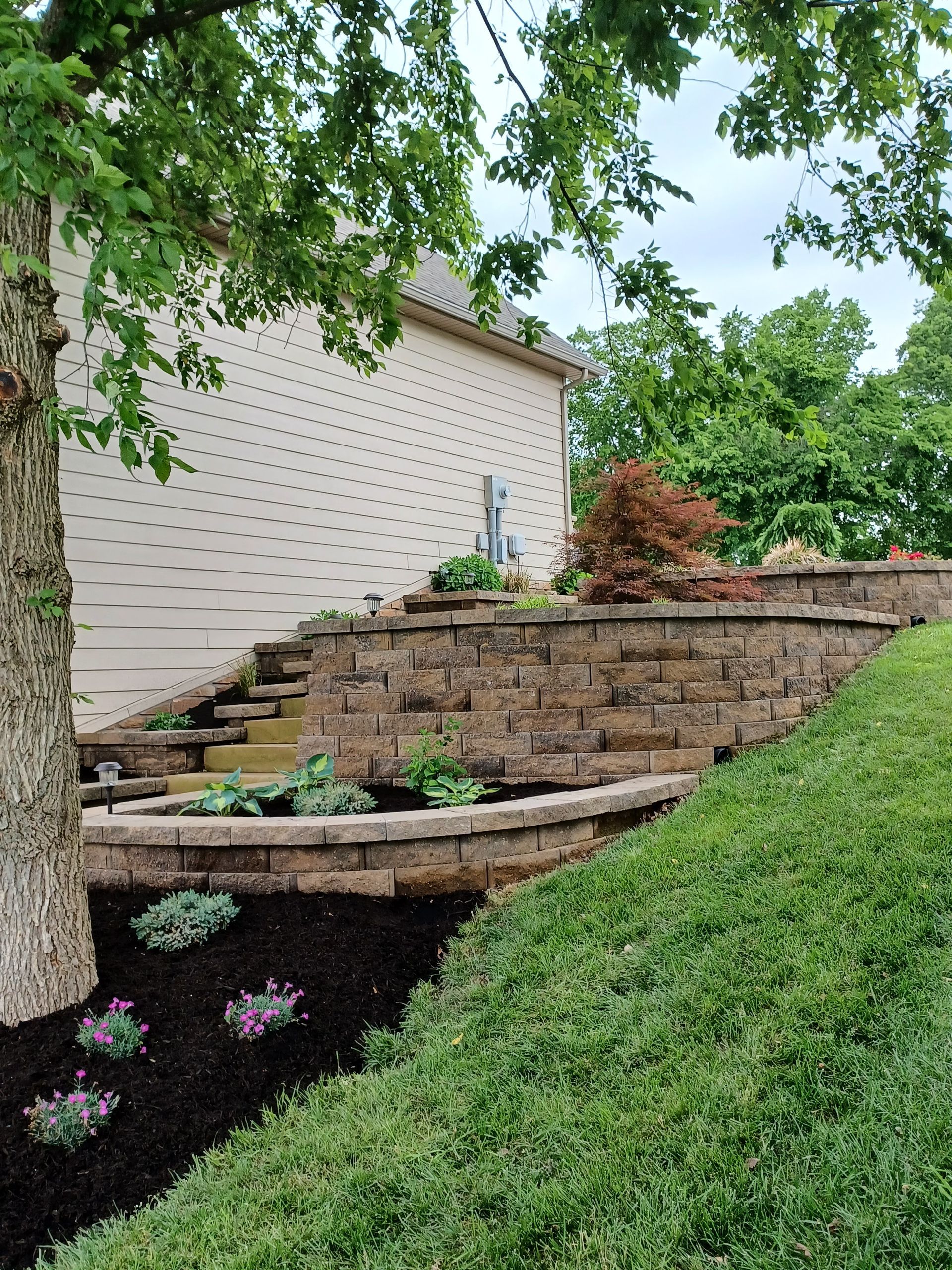 Landscaped tiered retaining wall with plants and a house in the background.