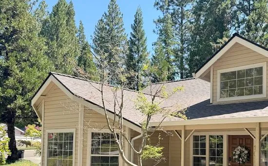 A beige, one-story house with a brown roof and a front porch, set against a backdrop of tall pine trees.