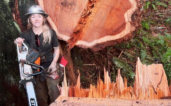 Woman with a chainsaw stands next to a freshly cut tree trunk in a forest.