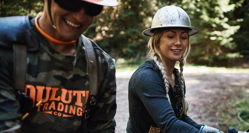 Man and woman smiling outdoors, wearing hard hats and work clothes, Duluth Trading logo visible.
