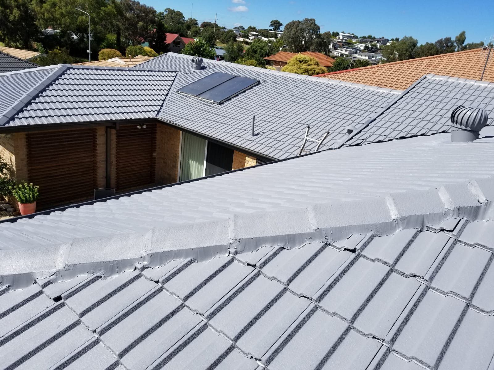 Gray tiled roof with solar panels and a vent, under a blue sky with houses in the background.