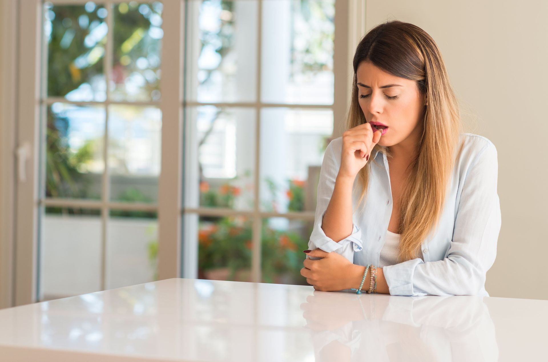 Woman indoors, coughing, resting hand on chin, eyes closed, sitting at a white table.
