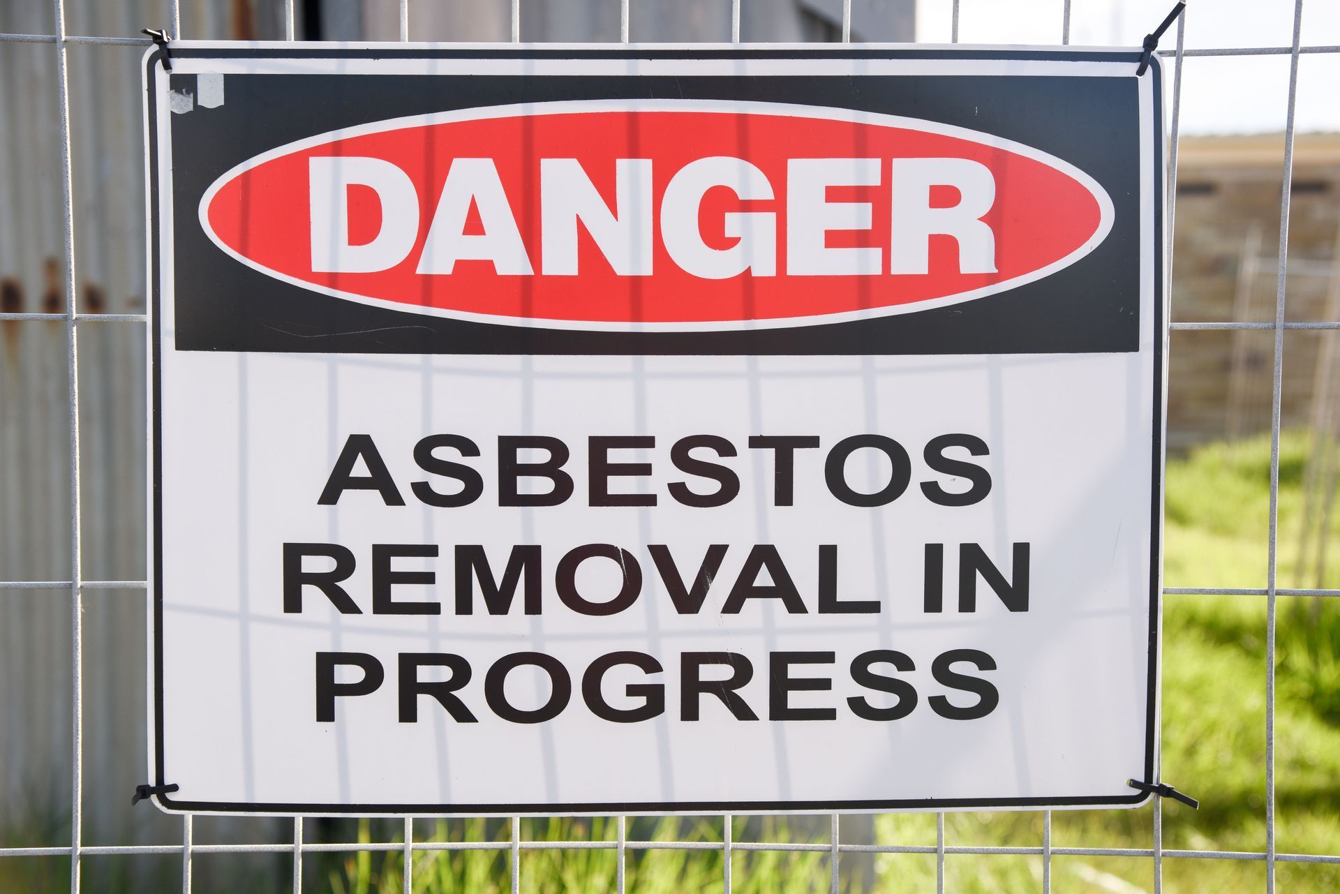 A danger sign on a chain-link fence warns of asbestos removal in progress. The sign is white, black, and red.