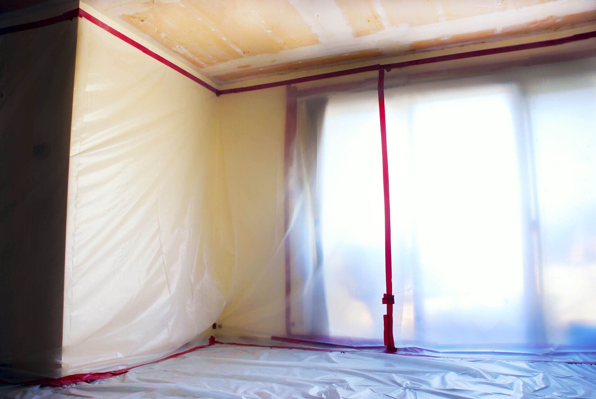 A room sealed with plastic sheeting and red tape, likely for renovation or abatement. Sunlight streams in from a window.