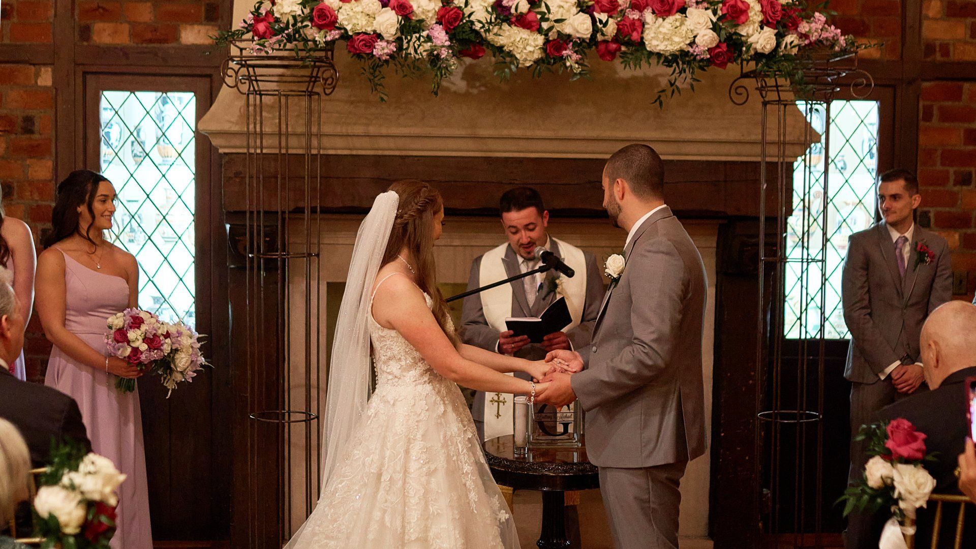 A bride and groom are holding hands during their wedding ceremony in front of a fireplace.