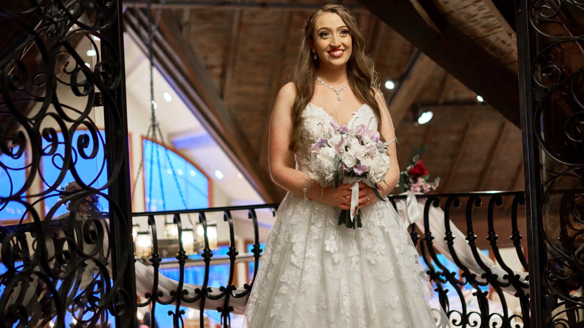 A bride in a wedding dress is standing on a balcony holding a bouquet of flowers.
