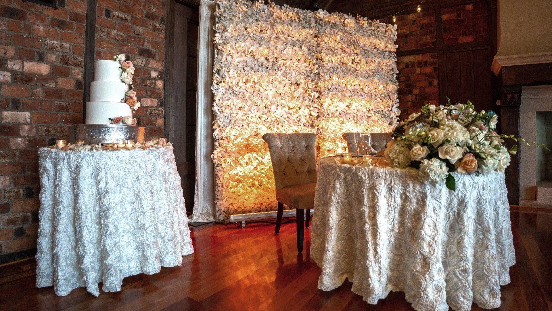 A wedding cake is on a table in front of a wall of flowers.