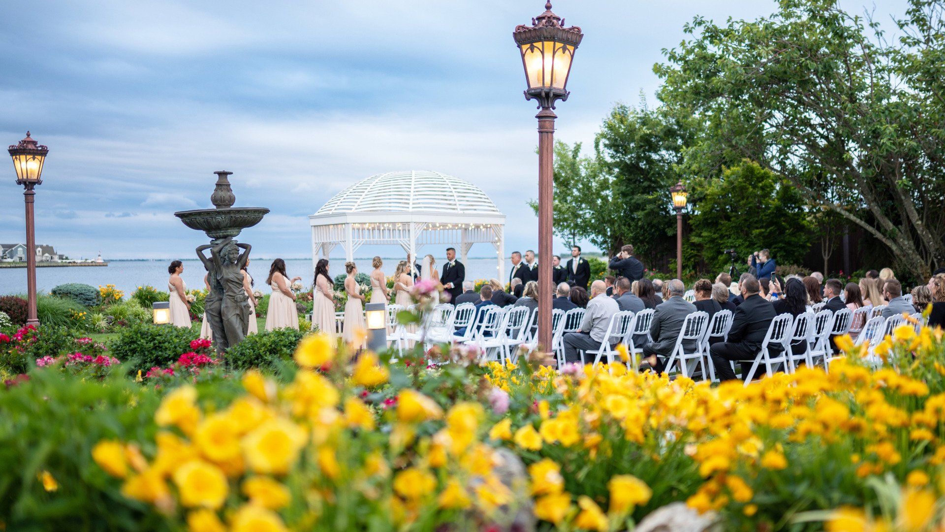 A wedding ceremony is taking place in a garden with yellow flowers and a fountain in the background.