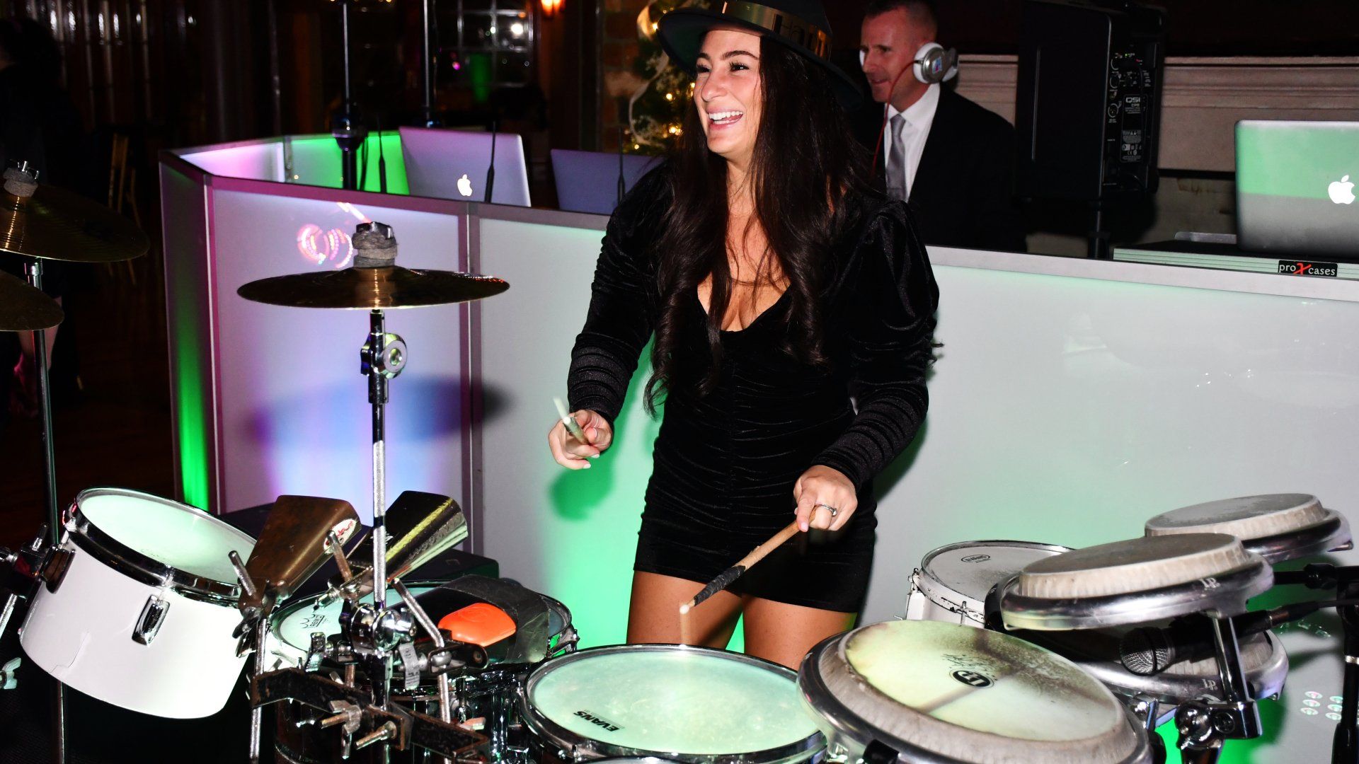 A woman in a black dress is playing drums in front of a dj.