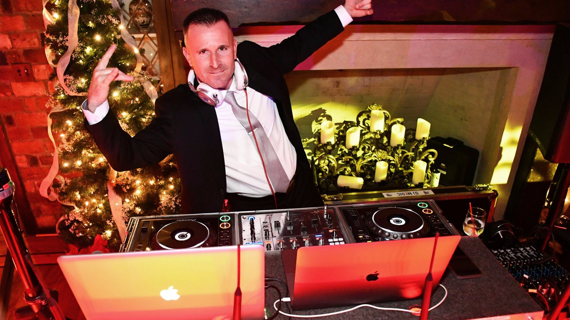 A man in a suit and tie is standing at a dj booth in front of a christmas tree.