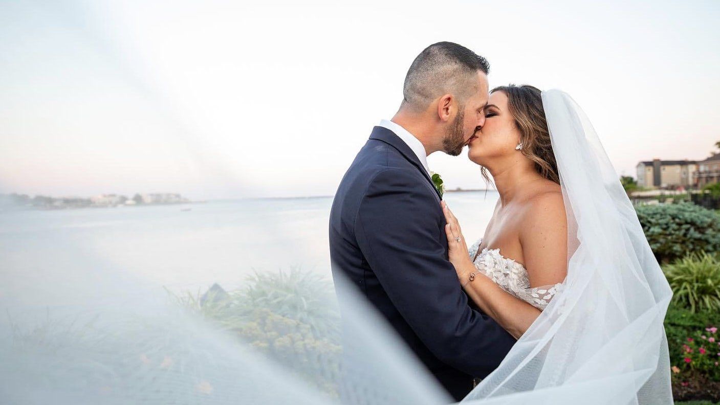 A bride and groom are kissing under a veil in front of a body of water.