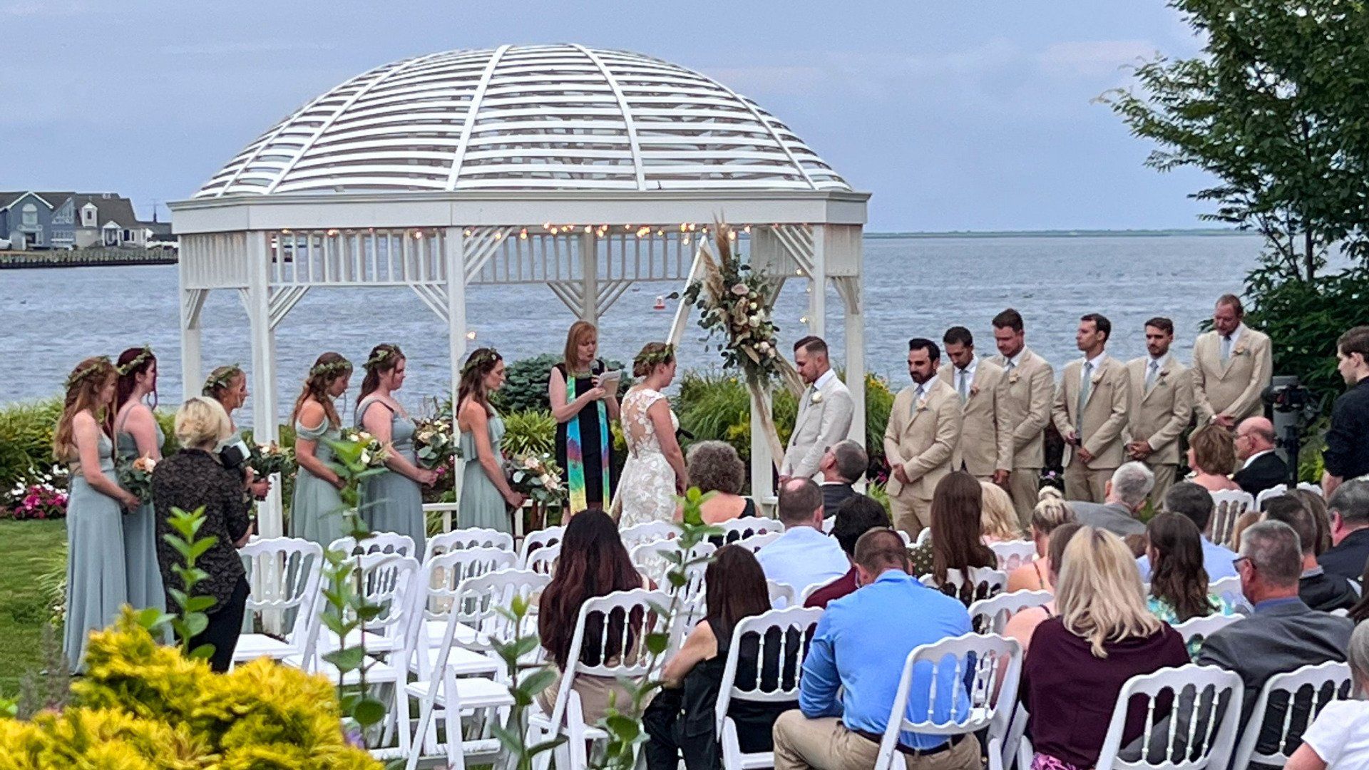 A wedding ceremony is taking place in front of a gazebo overlooking the ocean.