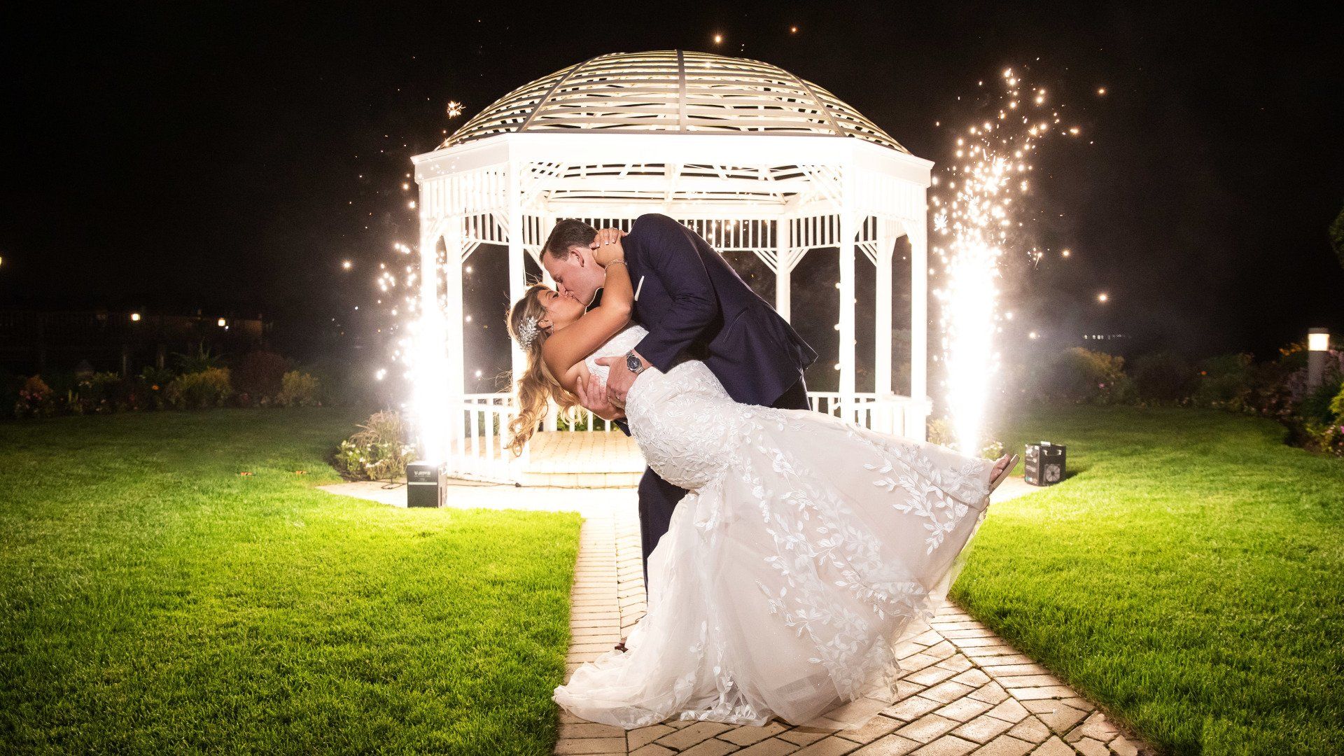 A bride and groom are kissing in front of a gazebo at night.