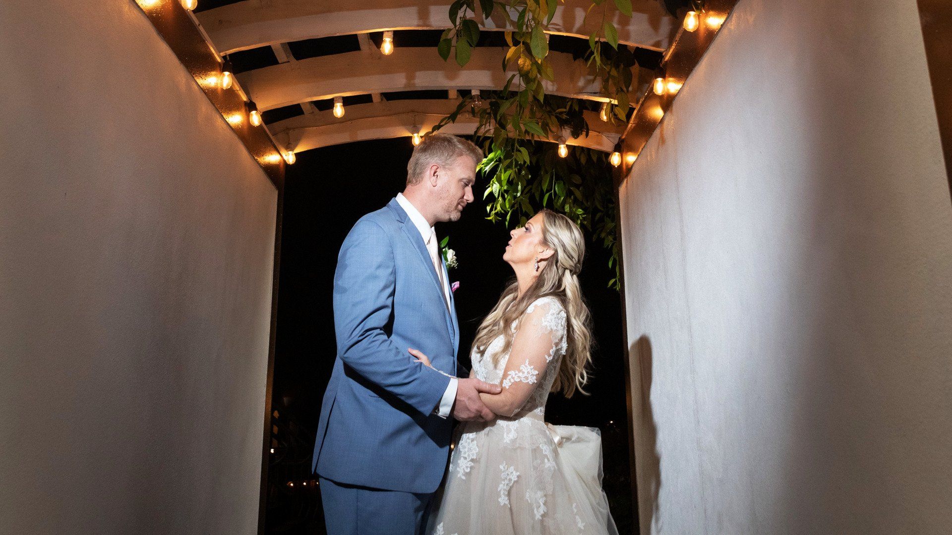 A bride and groom are standing next to each other under a pergola at night.