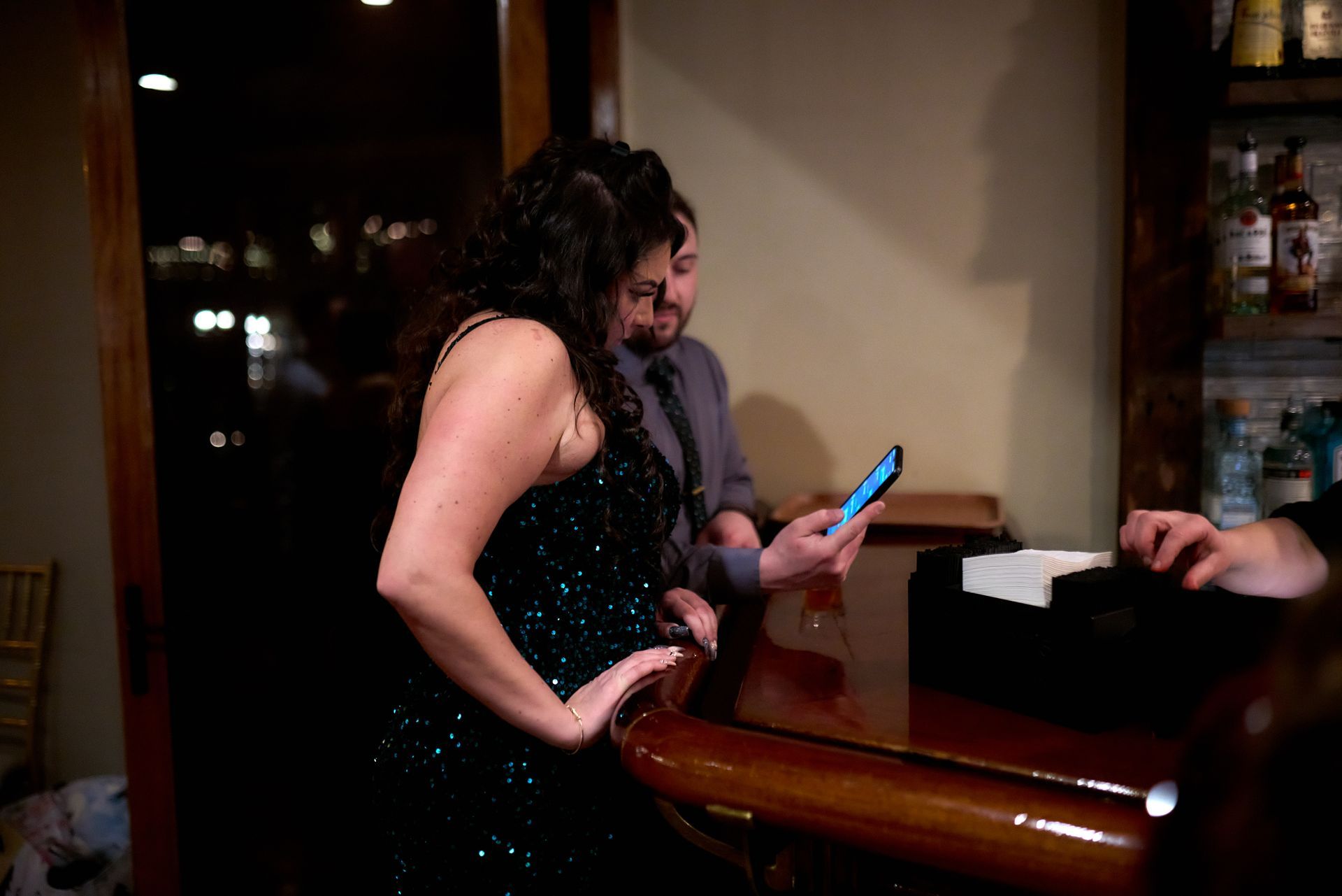 A woman in a black dress is standing at a bar looking at her phone.