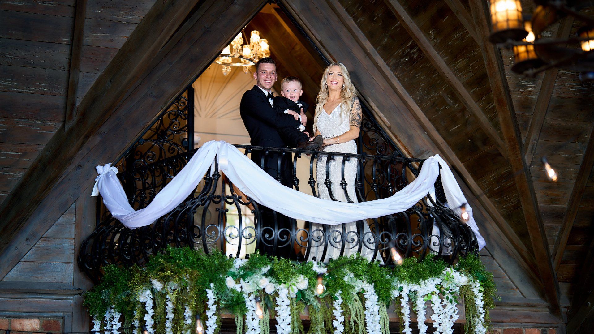 A bride and groom are standing on a balcony holding a baby.