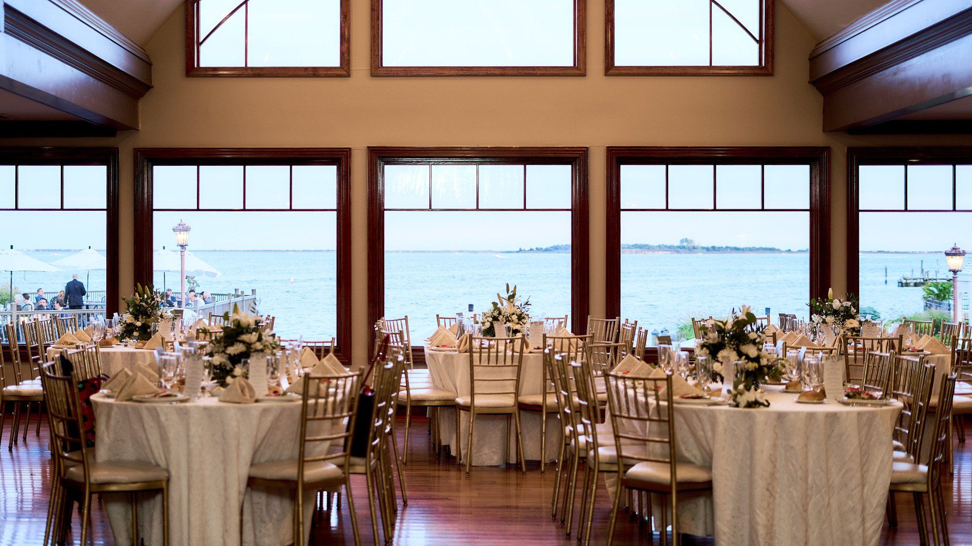 A large room with tables and chairs set up for a wedding reception with a view of the ocean.