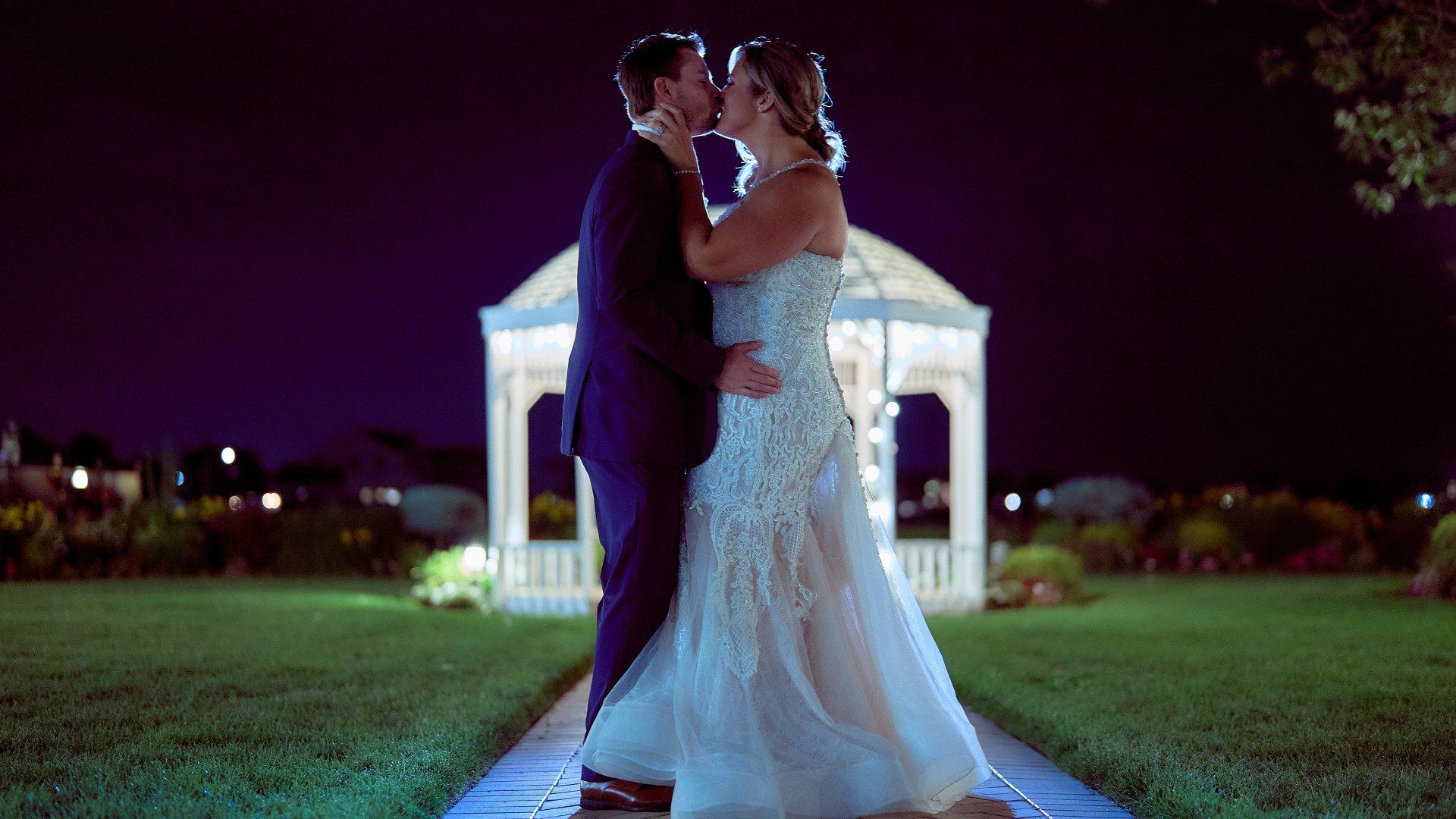 A bride and groom are kissing in front of a gazebo at night.