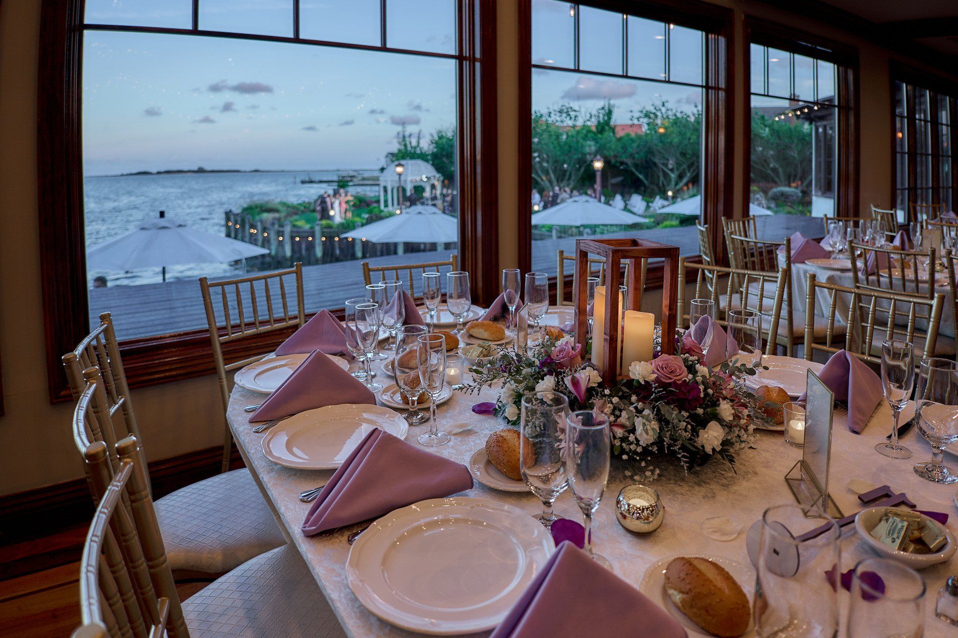 A long table with plates , glasses , napkins and candles in front of a window overlooking the ocean.